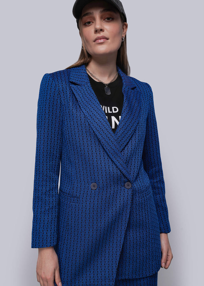 S=Woman wearing a blue textured blazer over a black outfit with visible text on a plain background