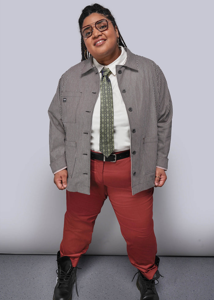 Person wearing a gray jacket, white shirt, red pants, and black shoes against a plain background