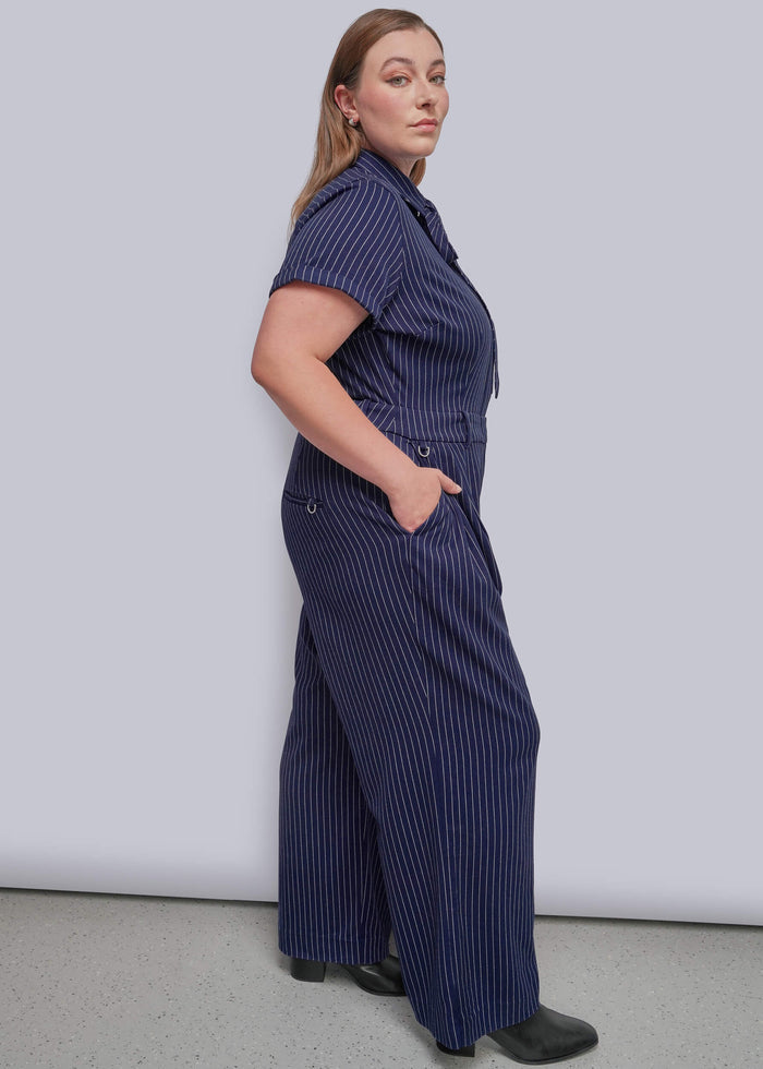 1X=A woman modeling a navy blue short-sleeve jumpsuit with thin white vertical pinstripes, standing against a light gray studio background.