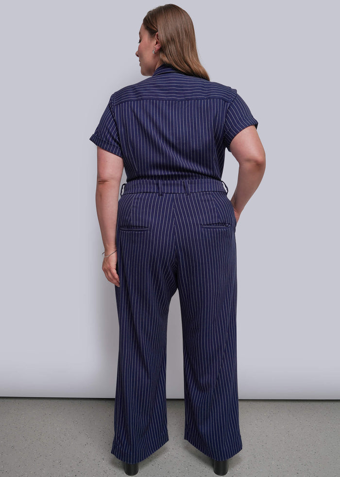 1X=A woman viewed from the back wearing a navy blue short-sleeve jumpsuit, standing against a plain light gray wall.