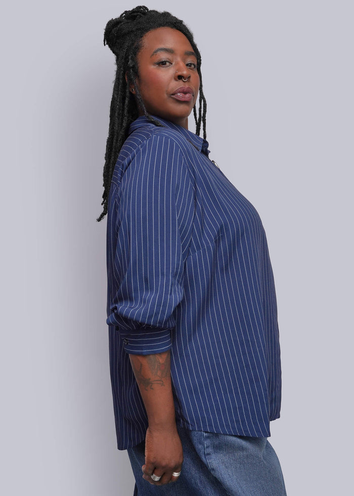 1X=A person with long black braids is shown from the side, wearing a blue and white vertically striped button-up shirt and blue denim pants against a plain light gray background.