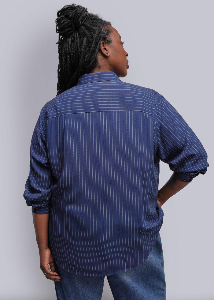 1X=A person shown from the back wearing a blue button-up shirt with thin white vertical pinstripes and blue jeans, standing against a plain light gray studio background.
