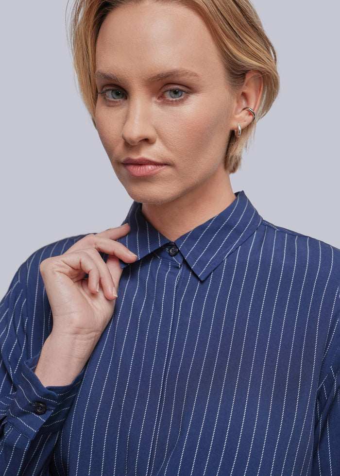S=A close-up of a woman wearing a blue button-down shirt with thin white vertical pinstripes, set against a solid light gray background.