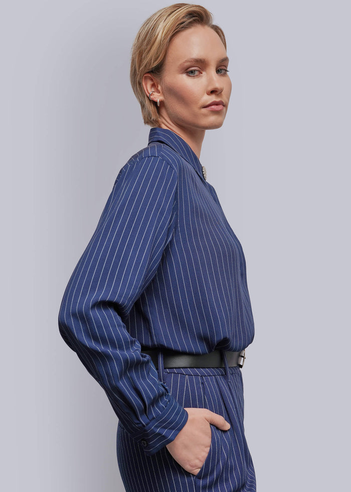 S=A woman wearing a navy blue pinstriped long-sleeve shirt and matching trousers, styled with a black belt, photographed against a solid light gray background.