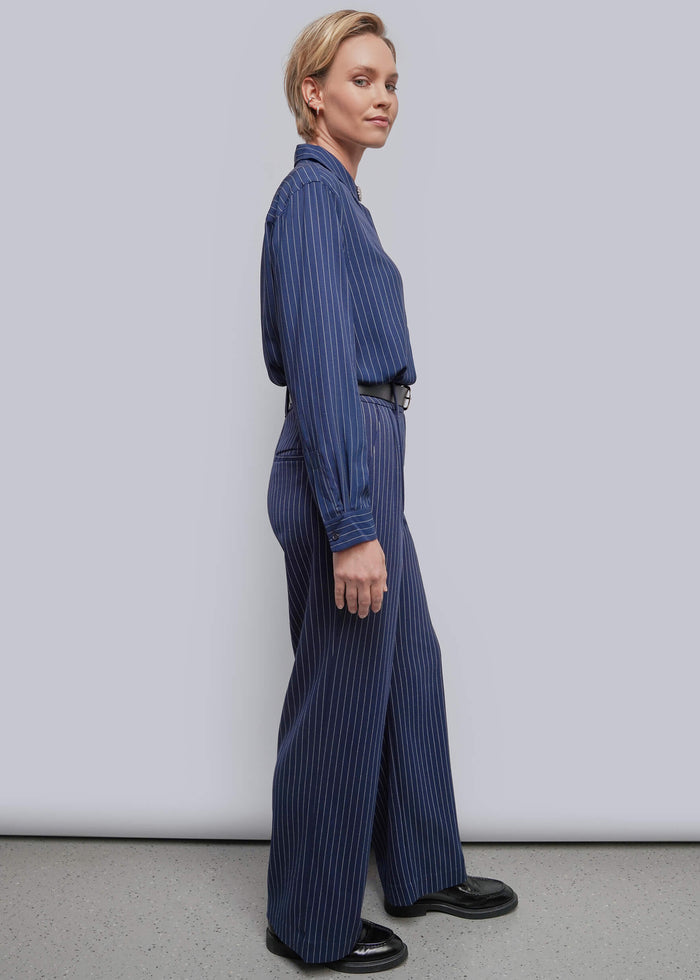 S=A woman stands in profile wearing a blue pinstriped long-sleeve shirt and matching wide-leg trousers with a black belt, against a plain light gray studio background.