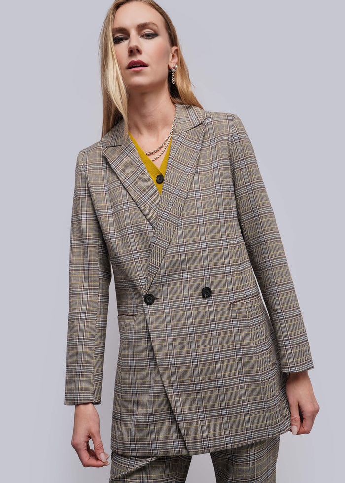 S=A woman wearing a double-breasted plaid blazer in beige, brown, and gray tones, styled with a yellow top and layered necklaces, against a solid gray studio background.