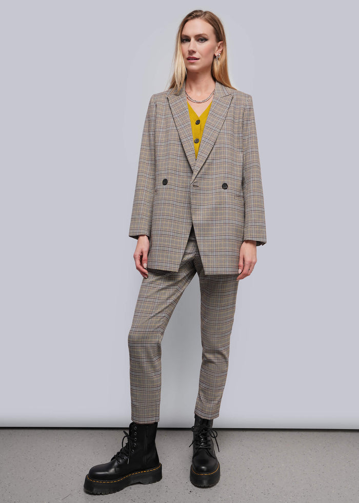 S=A woman modeling a beige and brown plaid double-breasted blazer with matching trousers, worn over a yellow top and paired with black combat boots, against a plain gray studio background.