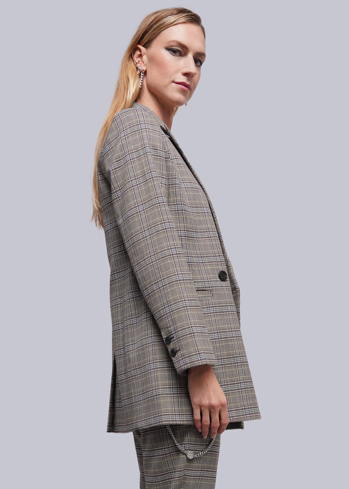 S=A woman modeling an oversized plaid blazer and matching trousers in a gray and beige check pattern, shown from a side profile against a solid gray studio background.