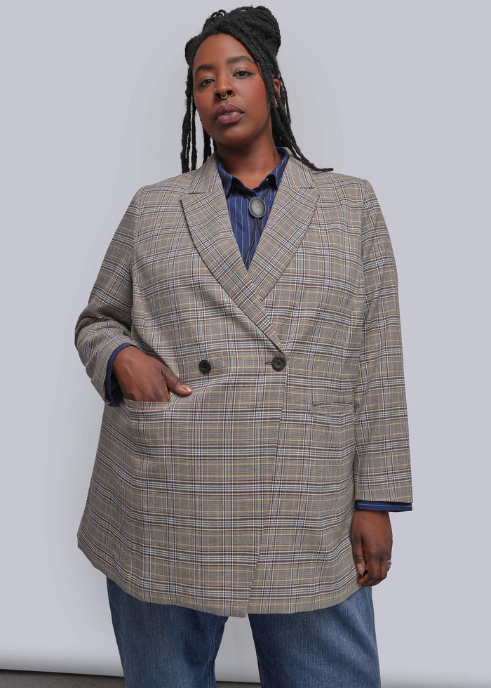 1X=A model wearing a beige and brown plaid double-breasted blazer over a blue collared shirt and blue jeans, standing against a light gray studio background.