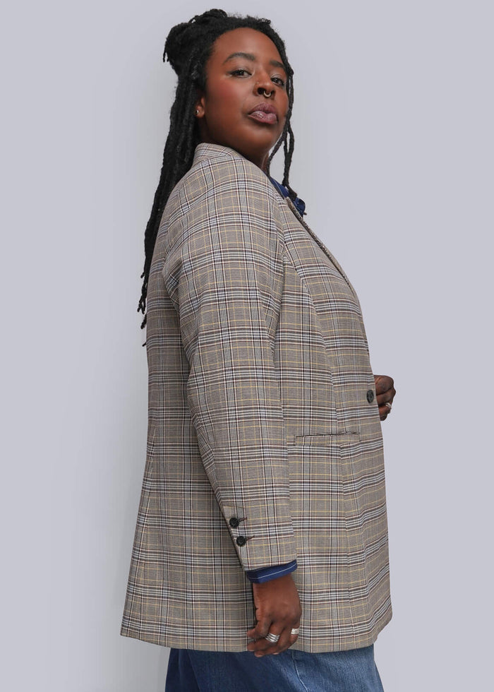 1X=A woman modeling a beige and brown plaid blazer with blue jeans and black ankle boots, standing against a plain light gray studio background.