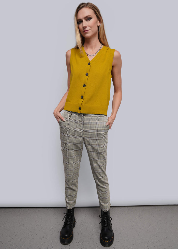 S=A woman modeling a sleeveless mustard yellow knit vest with black buttons, paired with beige plaid trousers and black boots, standing against a light gray studio background.