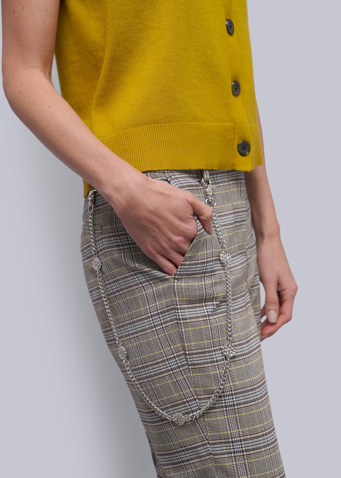 S=A close-up of a person wearing a mustard yellow sleeveless cardigan and beige plaid pants with silver chain details, against a light gray background.