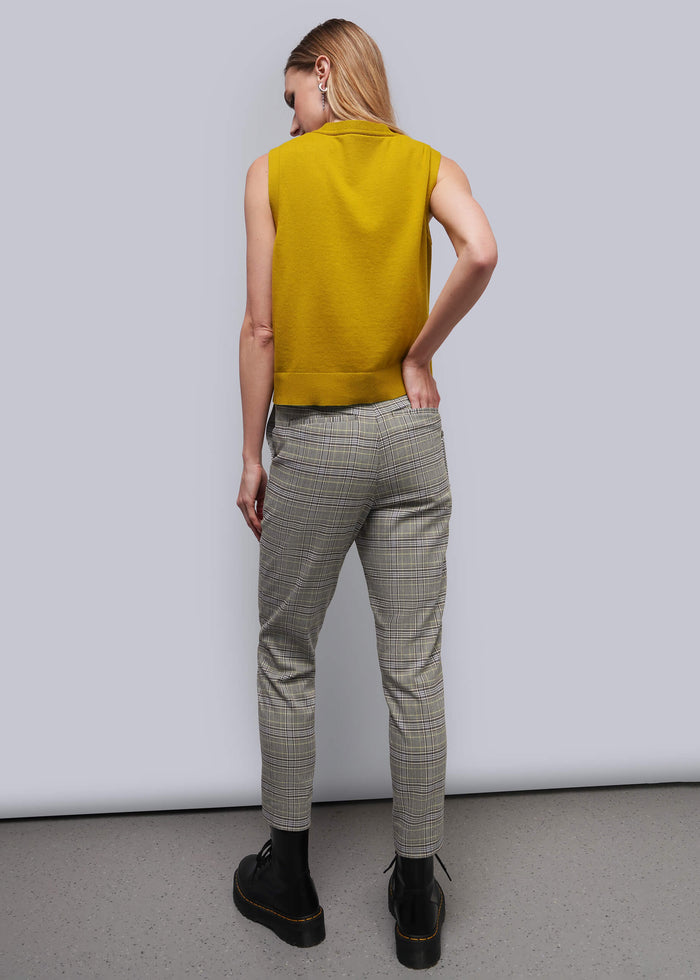 S=A woman shown from behind wearing a sleeveless mustard yellow knit top, gray and beige plaid trousers, and black boots against a light gray studio background.