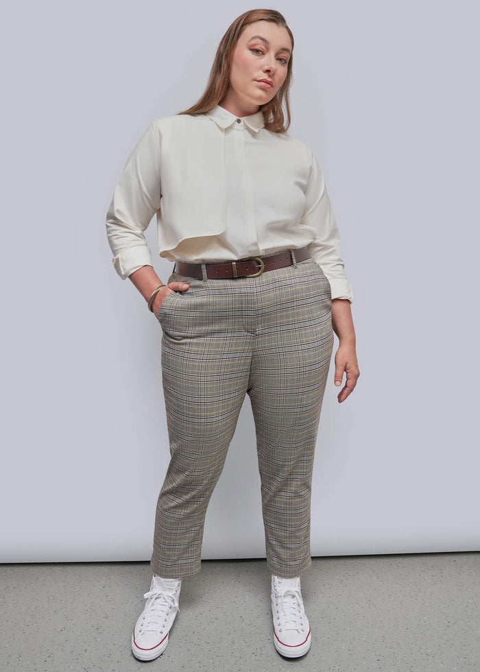 1X=A woman modeling a pair of beige and gray plaid trousers with a brown belt, paired with a cream shirt and white sneakers, standing against a light gray studio background.