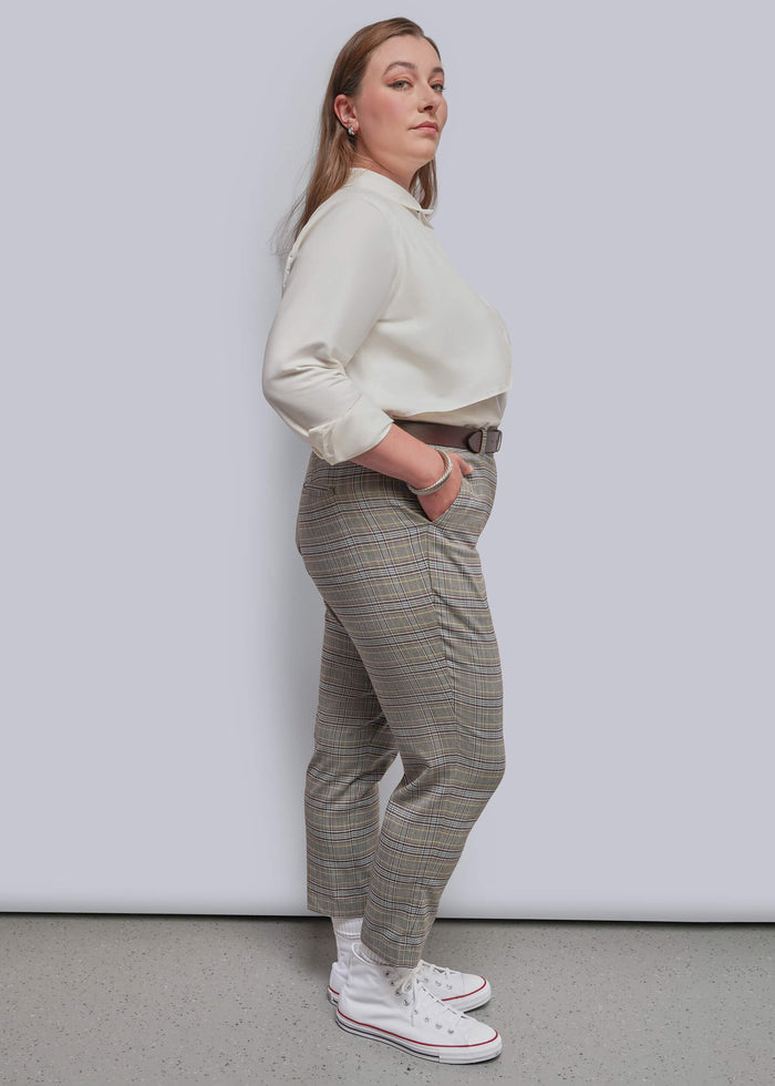 1X=A woman stands in profile wearing a cream long-sleeve top, gray and beige plaid trousers, white socks, and white sneakers against a light gray studio background.