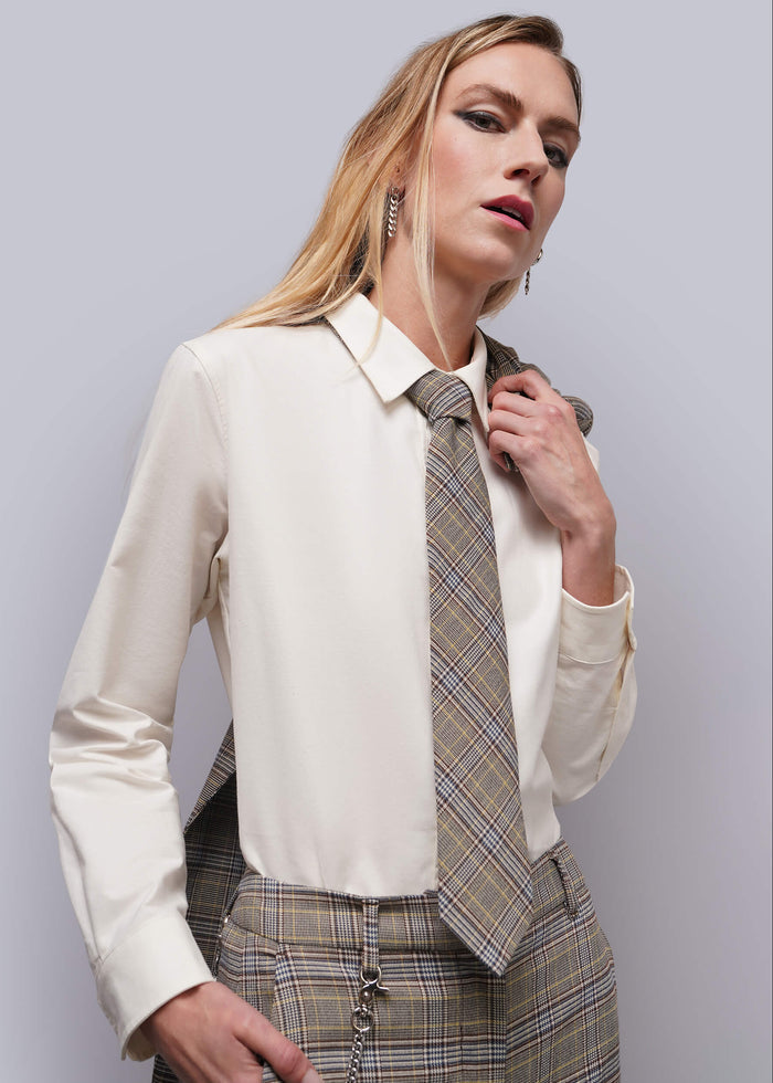 A woman modeling a cream long-sleeve blouse paired with a brown and beige plaid tie and matching plaid trousers, set against a solid light gray studio background.