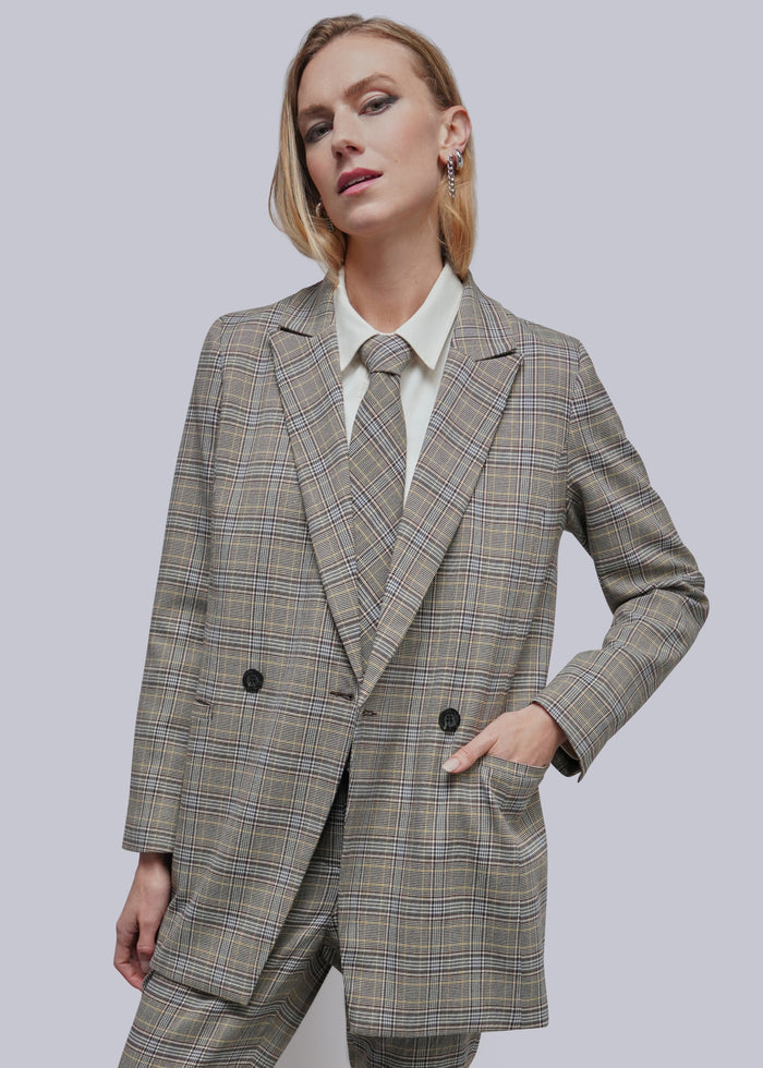 Model wearing grey plaid double breasted blazer and matching pant and tie against grey background.