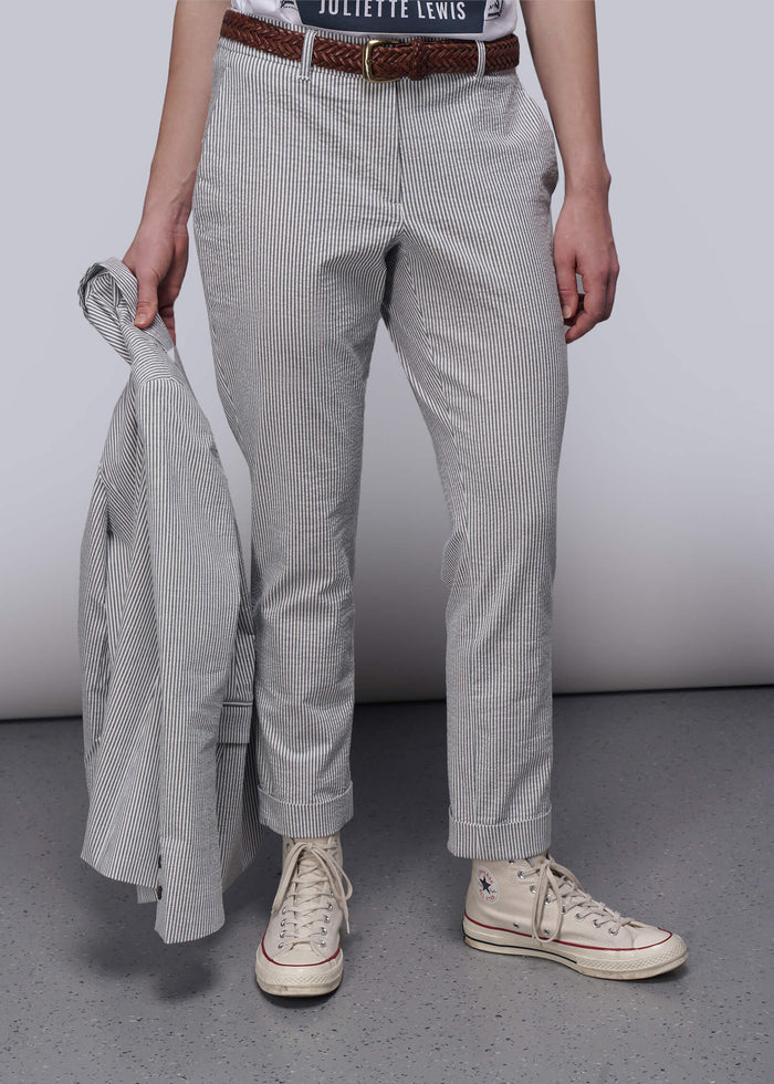 S=Person wearing light blue striped pants and white sneakers holding a gray jacket against a plain background