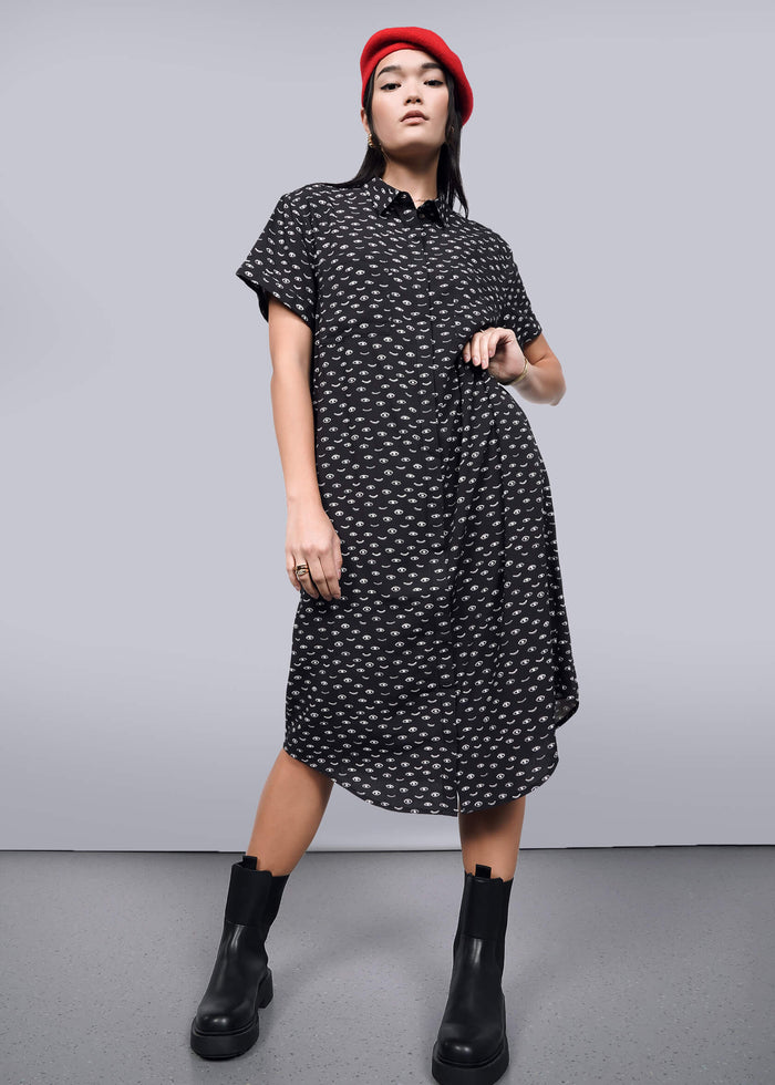 S= A woman in The Empower Shirt Dress, featuring a black pattern, button closure, and short sleeves below the knees, confidently poses with a red beret and black ankle boots. A plain background highlights her stylish outfit and poised stance.