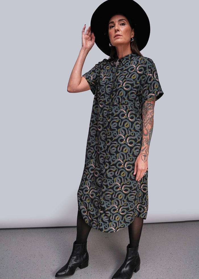 S=Woman wearing a patterned dress and black hat against a plain background