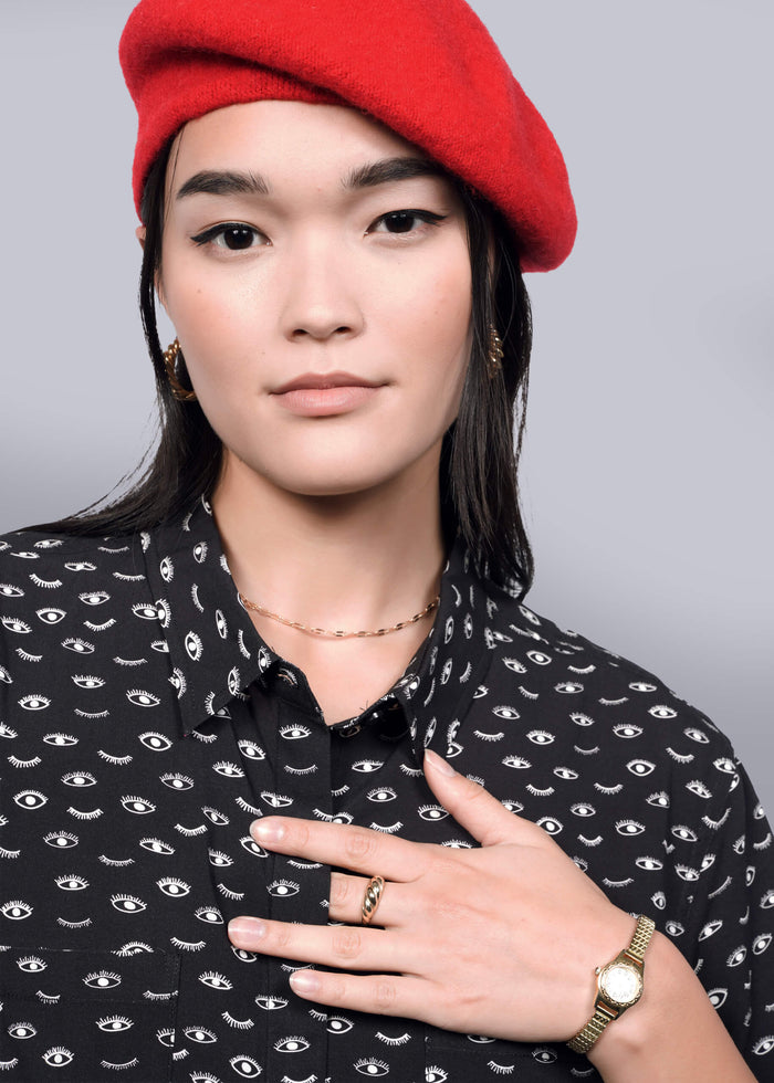 S= A person with long dark hair wears The Empower Shirt Dress, featuring eye patterns and a chic button closure, along with a red beret. They accessorize with gold hoop earrings, rings, and a watch. Their hand touches their chest against a white background.