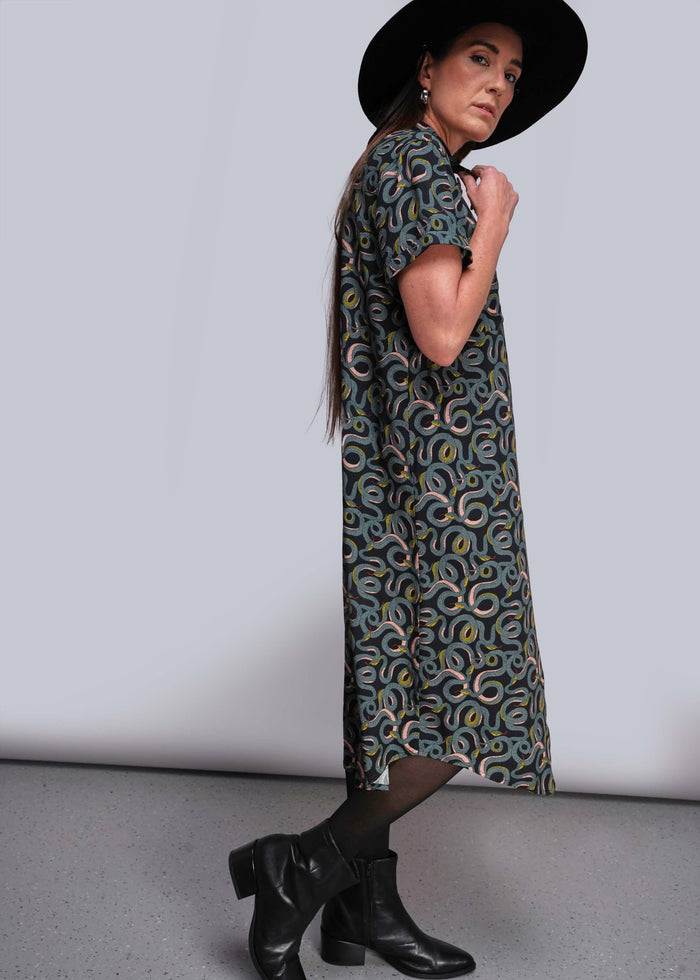 S=Woman wearing a patterned dress and black hat against a plain background