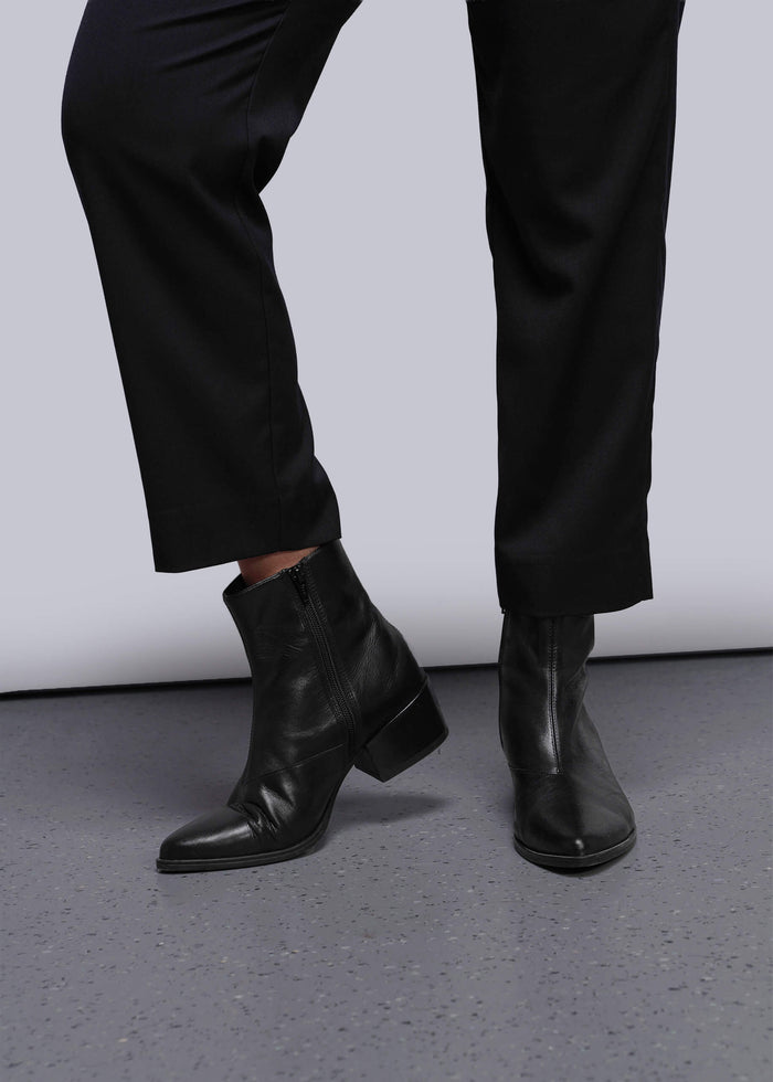 S=Black leather ankle boots worn with black pants on a gray floor.