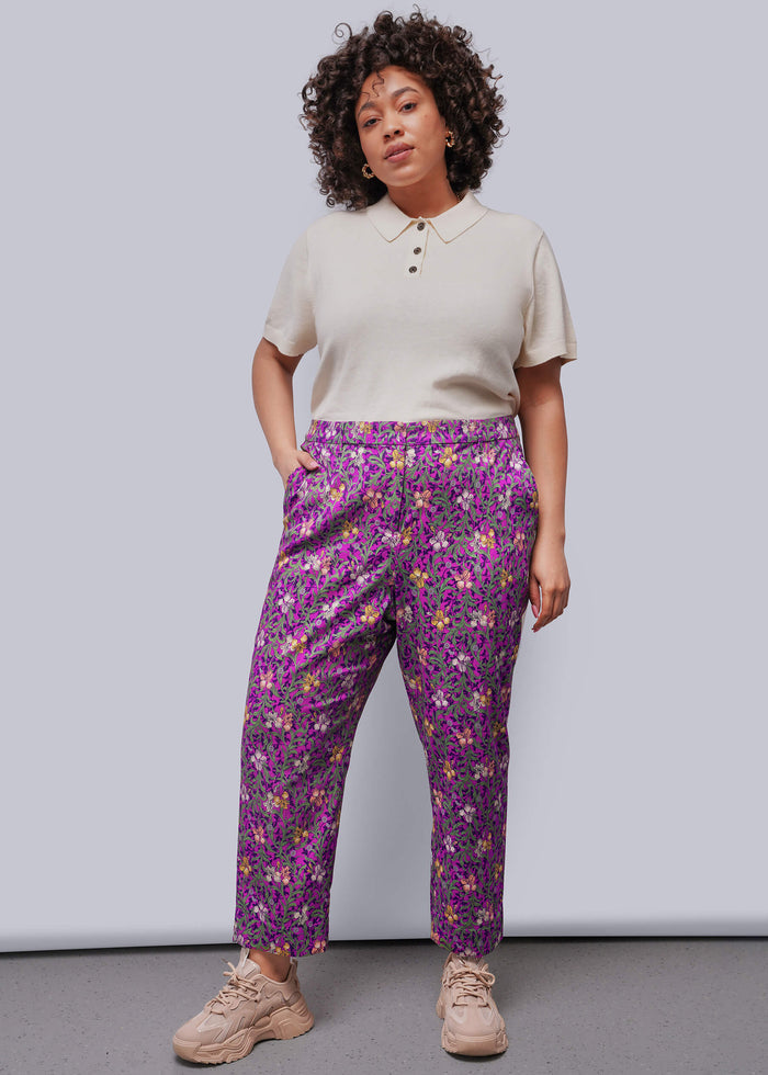 1X=Woman wearing a beige top and purple floral pants against a plain background