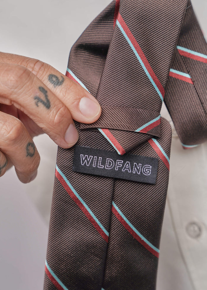 A close-up of a brown striped necktie with diagonal red and light blue lines, held by a tattooed hand. The brand label 'WILDFANG' is clearly visible on the back of the tie.