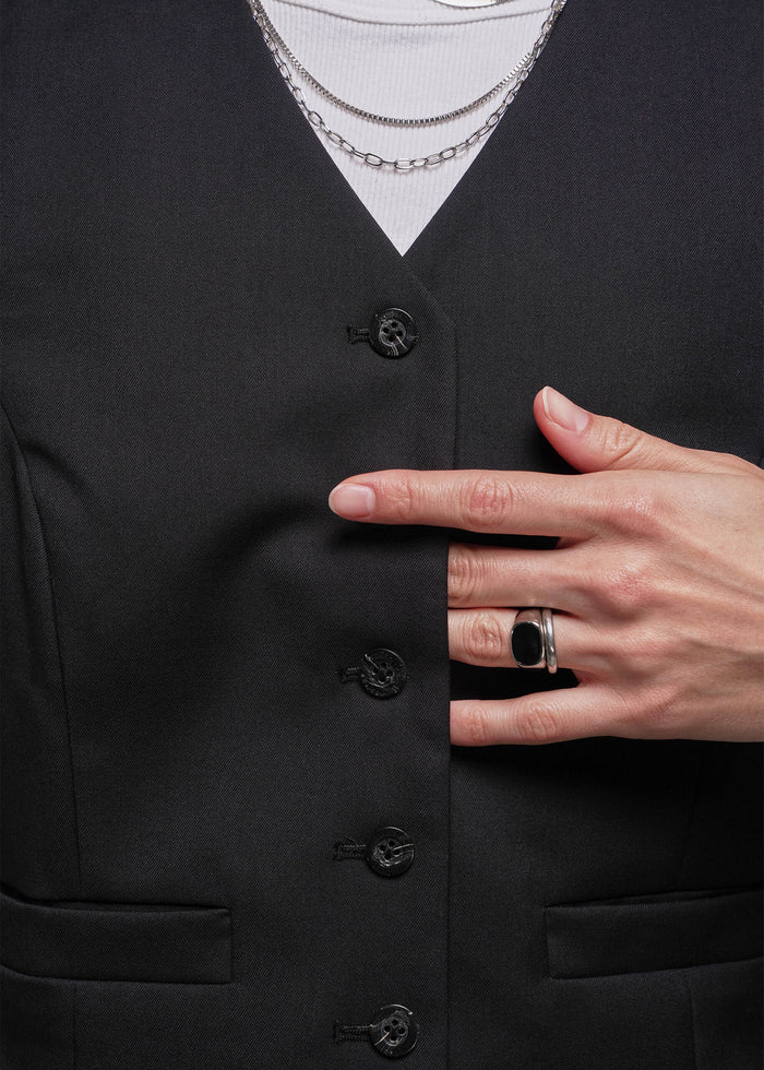 S=Close up of person wearing a black vest with buttons, white shirt, and layered necklaces.