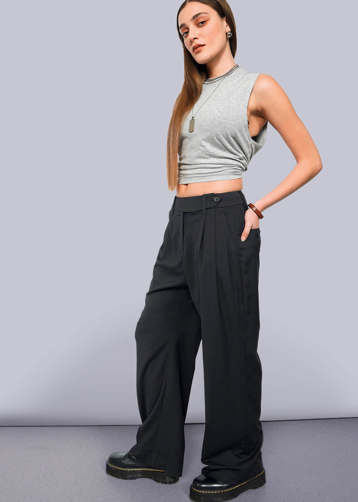 S=A woman with straight, long hair and a neutral expression stands against a white background, wearing The Empower Wide Leg Trouser with a sleeveless grey crop top, black platform shoes, and layered necklaces.