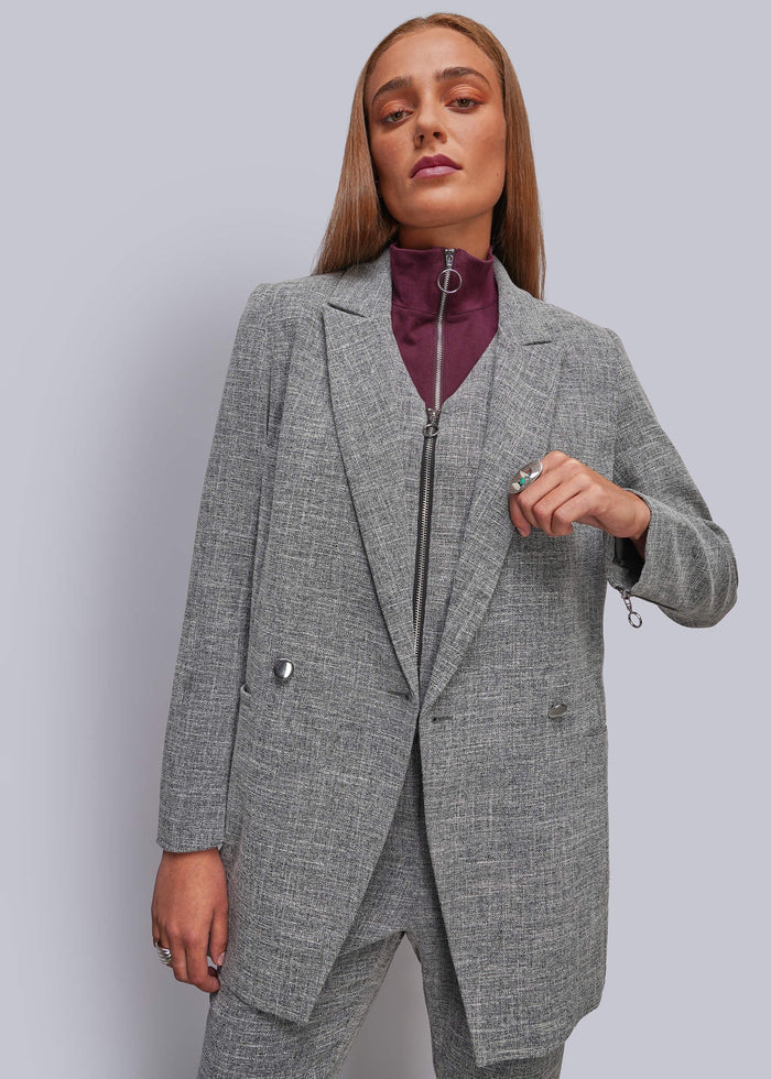S=A woman modeling a gray textured blazer and matching trousers with a zippered waist, worn over a dark purple high-neck top, against a solid light gray background.