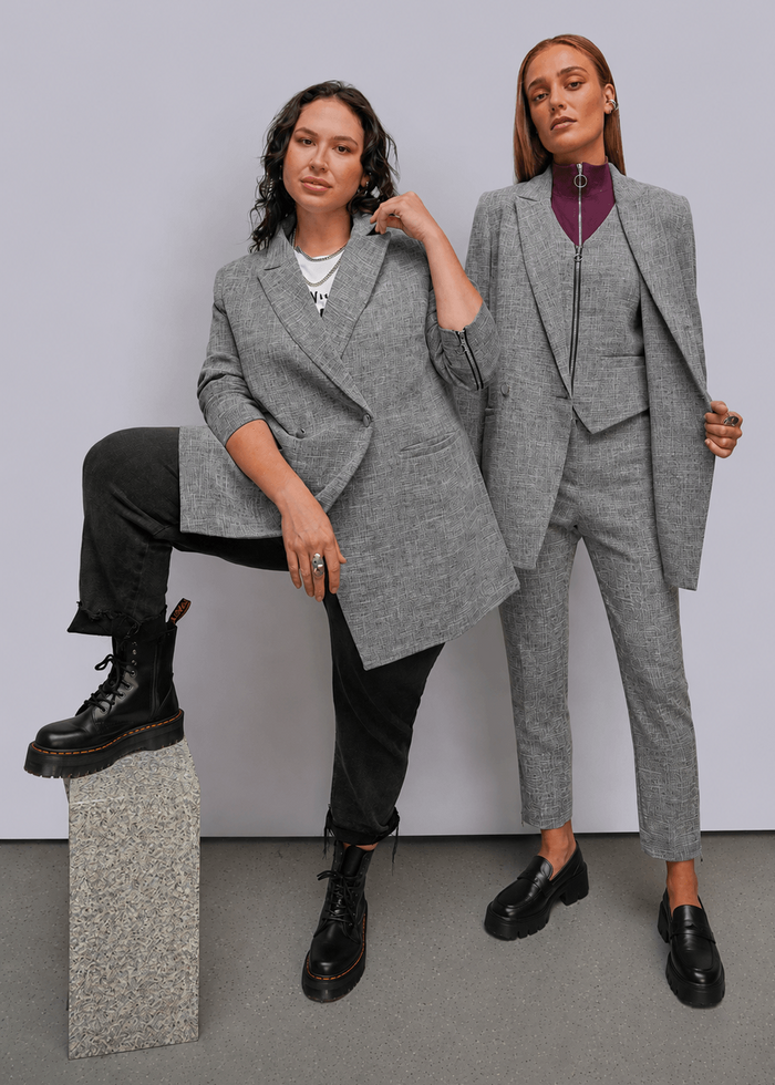 S=Two models posing in gray tailored suits against a light gray studio background. The model on the left wears an oversized gray blazer with black pants and boots, sitting on a stone block.