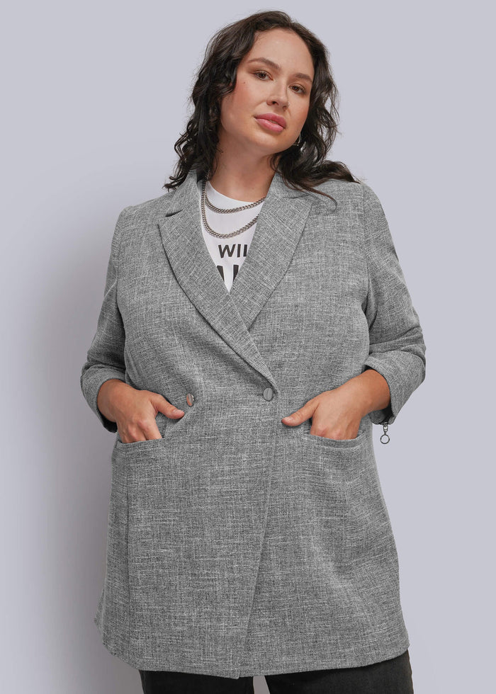 1X=A woman modeling a gray textured double breasted blazer with notch lapels and front pockets, worn over a white graphic t-shirt and black pants against a light gray studio background.