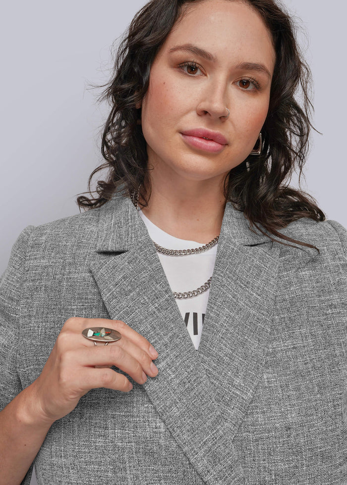 1X=A close-up of a woman wearing a textured gray blazer over a white t-shirt, accessorized with silver chain necklaces and a green ring.