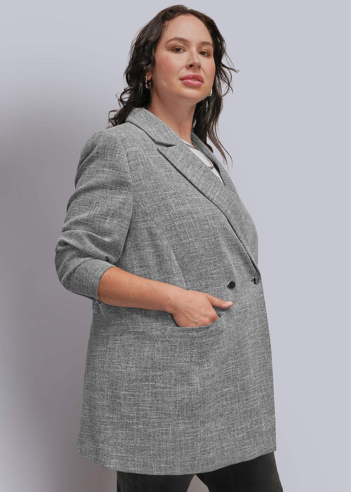 1X=A woman wearing a gray textured double-breasted blazer with black pants, standing against a light gray studio background.