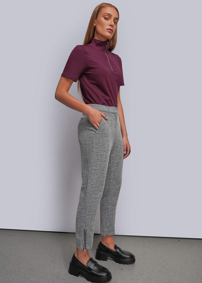 S=A woman modeling a short-sleeve burgundy mock-neck zip-up top and high-waisted gray textured ankle-length trousers with zipper details at the cuffs, paired with black loafers, against a light gray studio background.