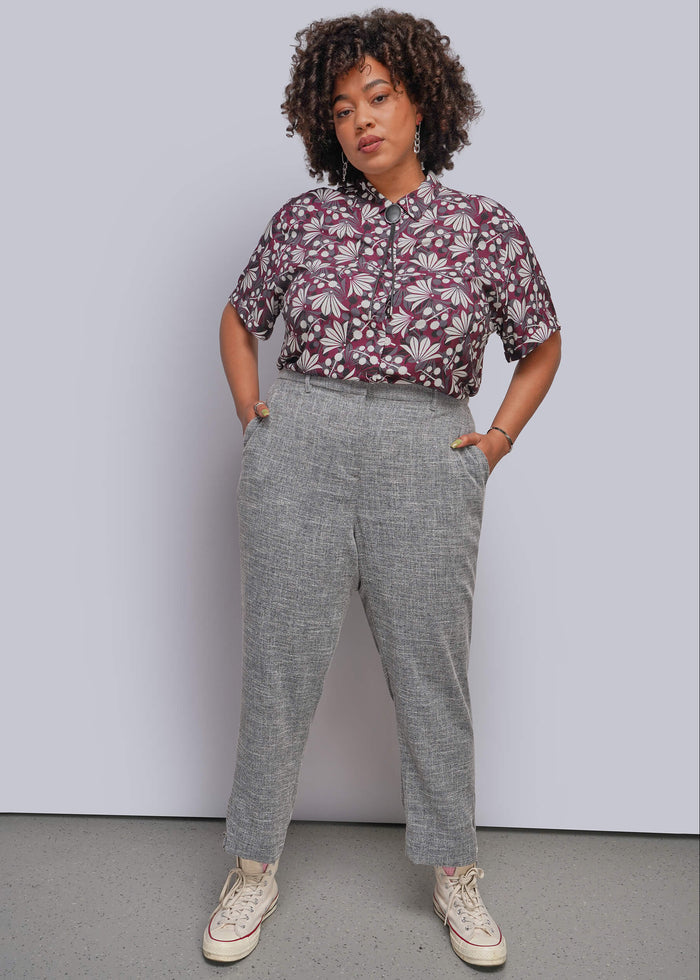 1X=A model stands against a light gray studio wall wearing a short-sleeve button-up shirt with a burgundy and white floral pattern, paired with heather gray cropped trousers and cream sneakers.