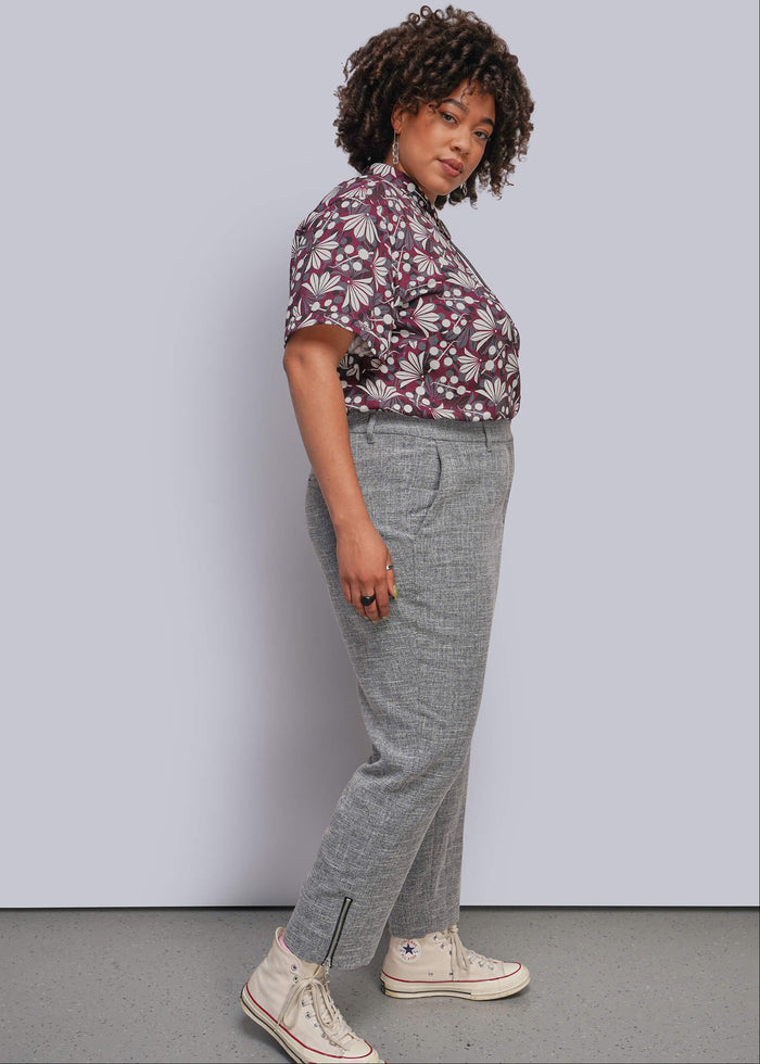 1X=A woman modeling a short-sleeve button-up shirt with a burgundy and white leaf pattern, paired with gray cropped pants and off-white high-top sneakers, standing against a light gray studio wall.
