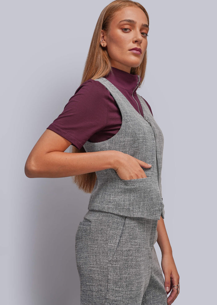 S=A woman modeling a gray two-piece vest and trouser set over a deep purple mock-neck t-shirt, photographed against a solid light gray studio background.