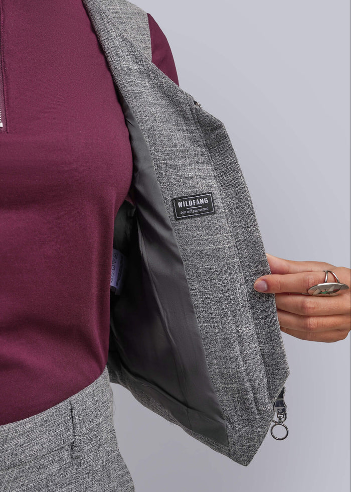 S=A close-up of a person wearing a gray textured vest over a burgundy top, with their hand holding open the vest to show the dark gray interior lining. A small black brand label reading 'WILDFANG' is visible on the blazer.