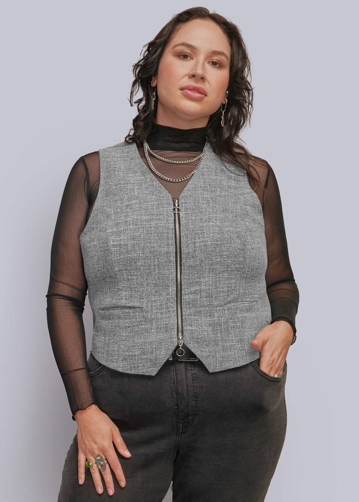 1X=A person modeling a sleeveless gray textured zip-front vest over sheer black long-sleeve mesh top, paired with dark gray jeans and layered necklaces, against a light gray studio background.