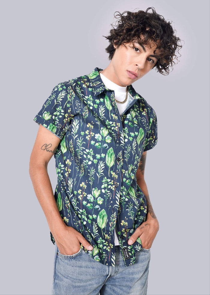 S= A person with curly hair confidently wears The Essential Button Up in navy with green floral patterns, tailored fit, and short sleeves. Underneath is a white shirt, paired with light blue jeans against a crisp plain white background. Hands are casually tucked in pockets.