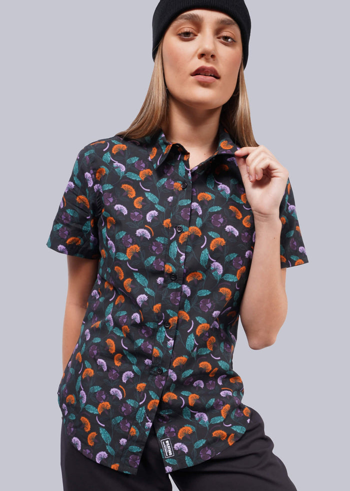 S= A person wearing a black beanie and The Essential Button Up, a short-sleeve shirt featuring a colorful mushroom pattern in breathable cotton, pairs it with tailored fit black pants while posing casually against a white backdrop.