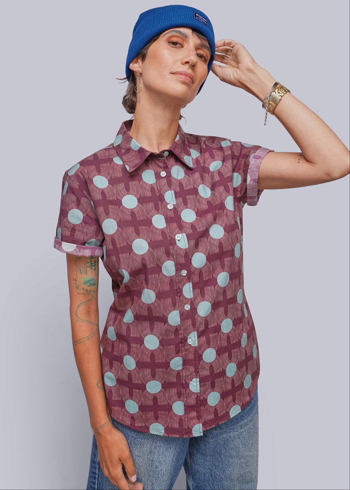 S=A person wearing a short-sleeve button-up shirt with a maroon and light blue geometric pattern, paired with blue jeans and a blue beanie, posed against a solid gray studio background.