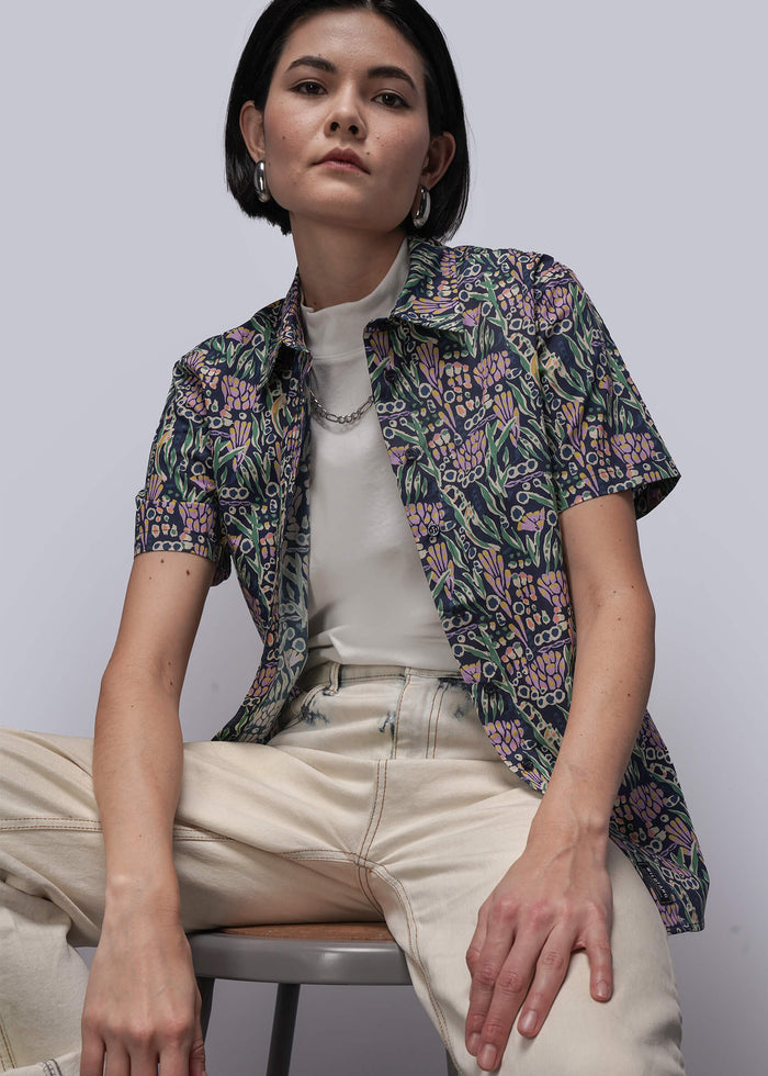 S=Woman sitting on a chair wearing a floral shirt over a white top and beige pants.