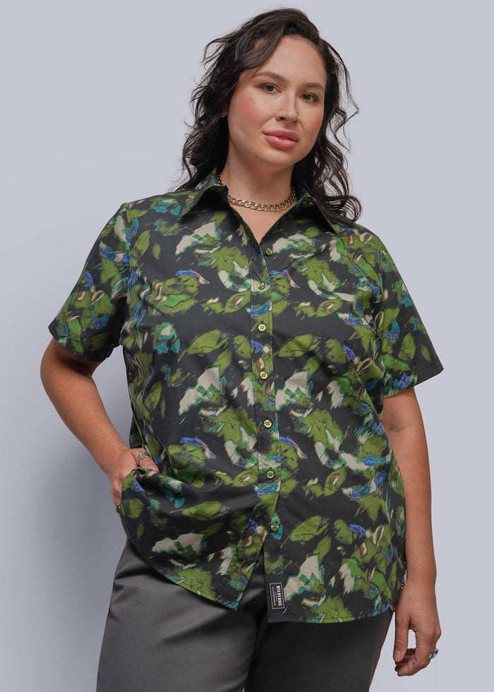1X=A woman modeling a short-sleeve button-up shirt with a green print on a black background, paired with gray pants, against a solid light gray studio background.