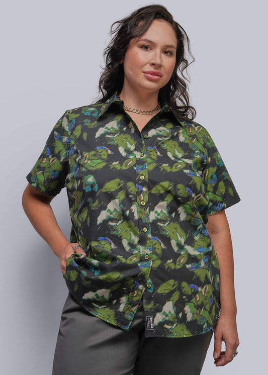 1X=A woman modeling a short-sleeve button-up shirt with a green print on a black background, paired with gray pants, against a solid light gray studio background.