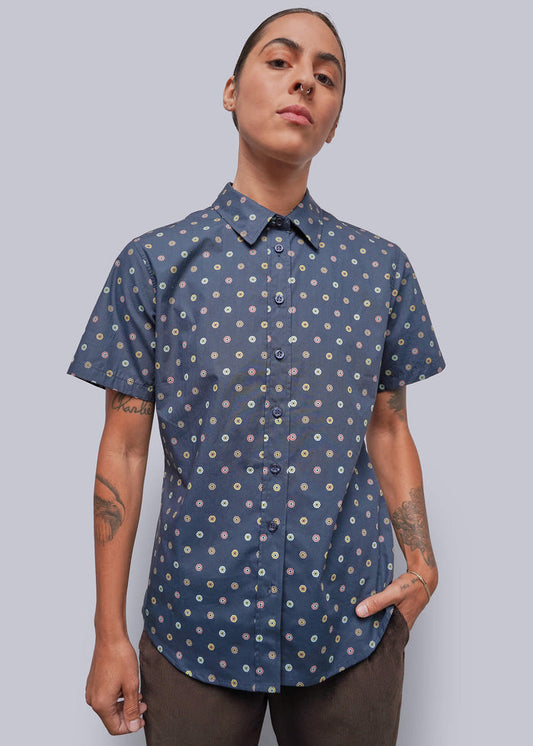 S=A model wearing a short-sleeve button-up shirt with a blue base and a small white and yellow floral pattern, paired with brown pants, against a solid gray background.