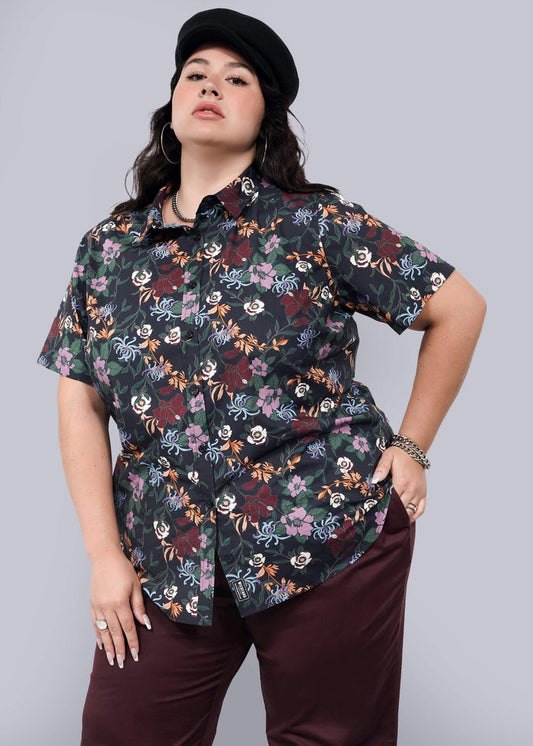 1X= A woman poses confidently against a plain light background wearing The Essential Button Up—a black floral short-sleeve, tailored-fit shirt with no chest gap—paired with dark burgundy pants, a black cap, and silver jewelry.